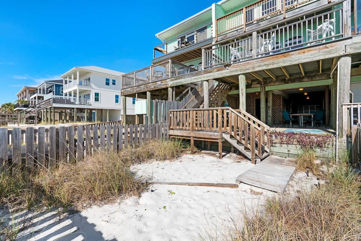 The back of a multi-level beach house with light green siding and wooden decks, featuring stairs leading down to a sandy area with a wooden fence and beach access.