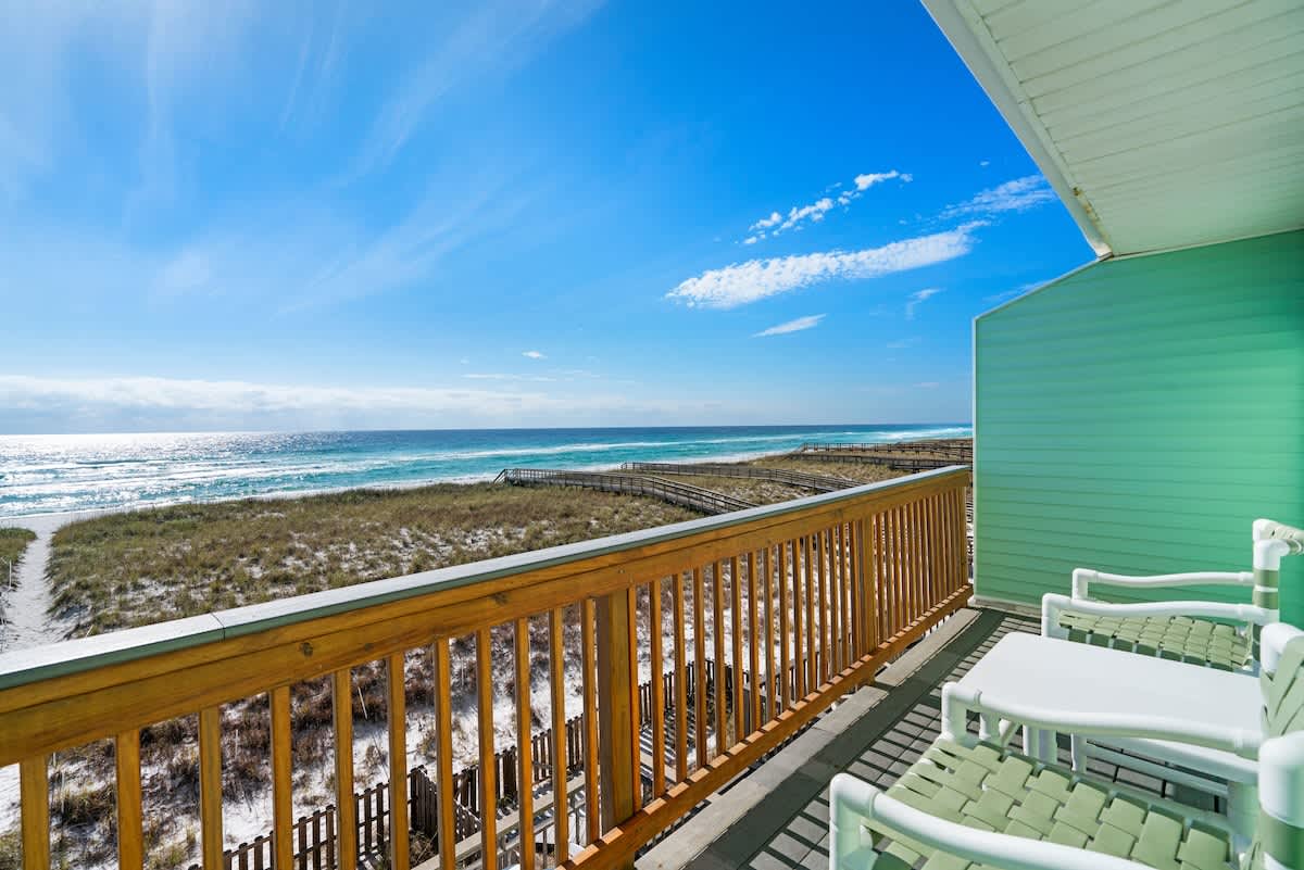 Expansive balcony overlooking the pristine sandy beach and the vast turquoise ocean, with a wooden railing and two white outdoor chairs. The vibrant blue sky completes the scenic view, perfect for enjoying the coastal breeze.