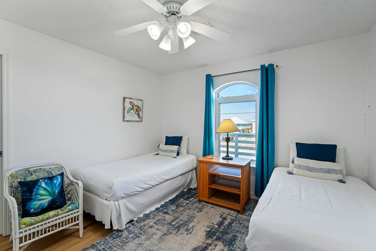 Second bedroom featuring two twin beds with white bedding, a white wicker chair, a wooden nightstand, and a window with teal curtains, decorated with coastal-themed art.