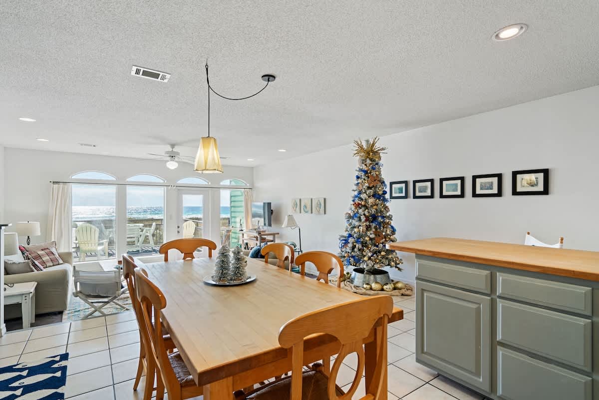 Open-concept dining area featuring a light wood dining table with six chairs on white tiled flooring, adjacent to a kitchen island and offering a glimpse of the living room and ocean view.