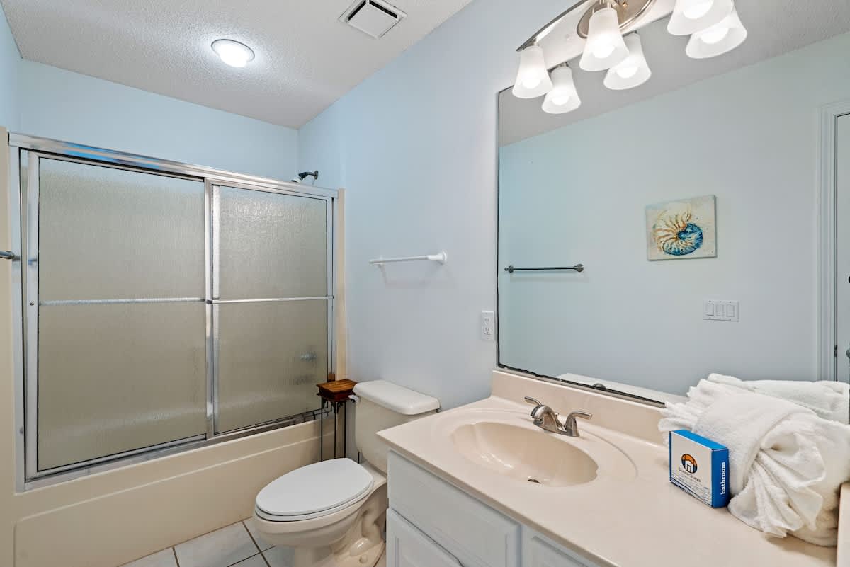 Light blue bathroom featuring a white vanity with a single sink, a toilet, and a shower-tub combo with frosted sliding glass doors. A seashell art piece hangs above the toilet, adding a coastal touch.