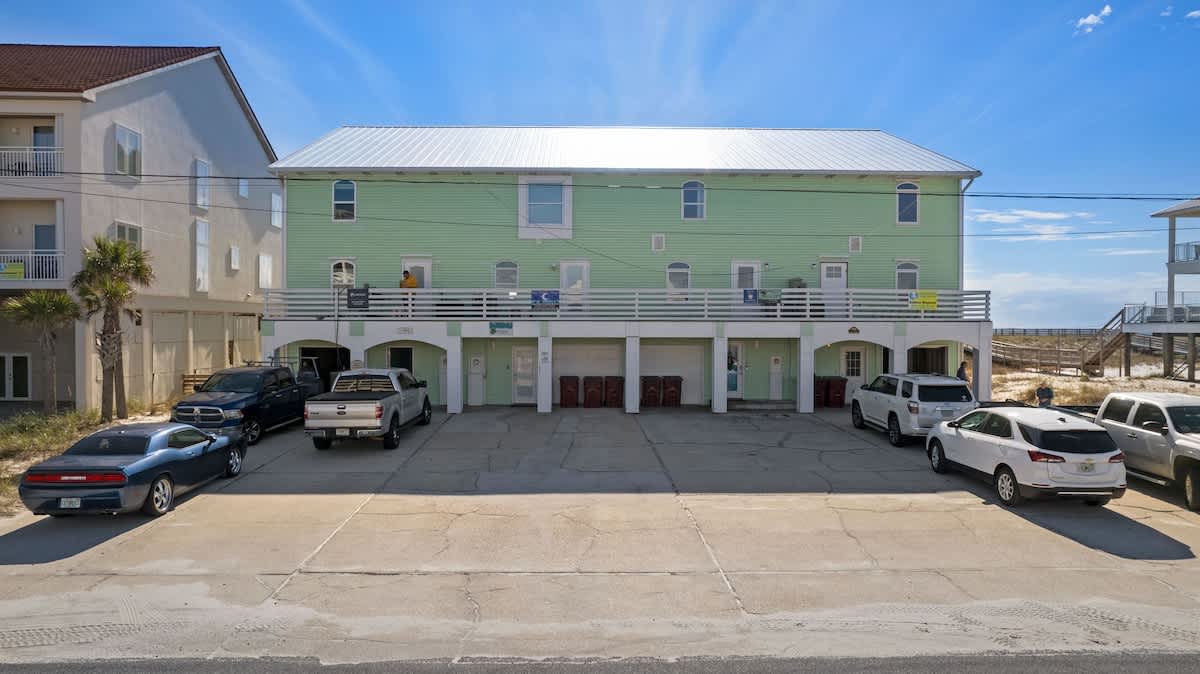 The front view of a light green, multi-story townhouse with a silver metal roof, featuring a ground-level parking area with several cars and a balcony on the second floor.