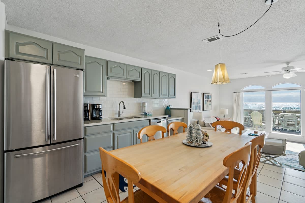 Modern kitchen with light blue shaker cabinets, white subway tile backsplash, stainless steel appliances, and a long wooden dining table with matching chairs, overlooking the ocean.