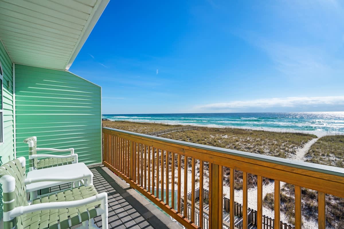 Balcony with a wooden railing, offering a stunning panoramic view of the sandy beach, dunes, and the turquoise ocean under a clear blue sky. Two white chairs and a small table are present for relaxation.
