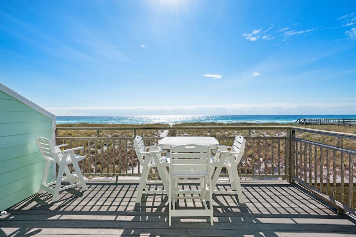 A wooden deck with light green siding on the left, furnished with a white plastic dining table and chairs, providing an unobstructed view of the sandy beach and the vast blue ocean.