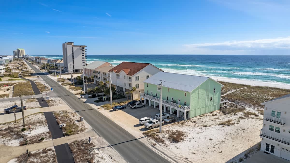 A wide aerial shot showcasing the light green townhouse and neighboring beach properties along a coastal road, with the expansive blue ocean and white sand beach stretching into the distance.