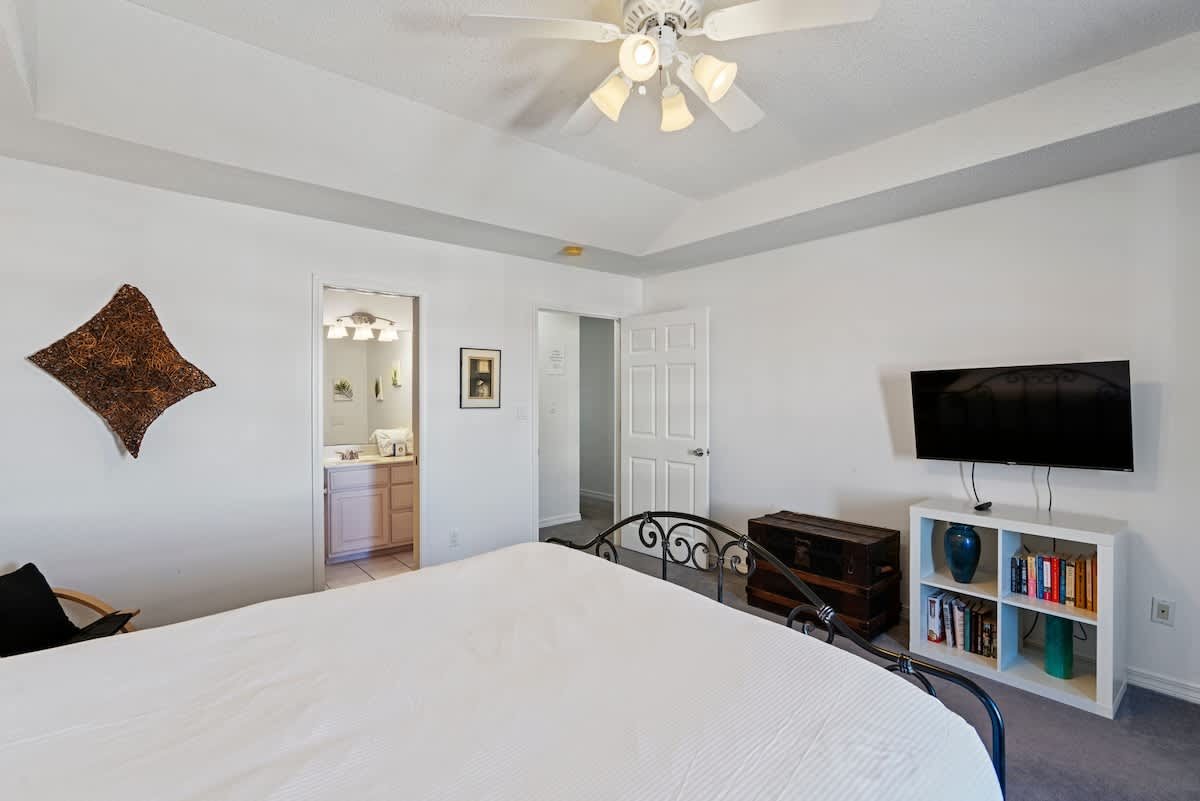 View of the bedroom showing the foot of the wrought iron queen bed, a wall-mounted TV above a white bookshelf filled with books, and a dark wooden trunk. An open doorway leads to a closet or another room.