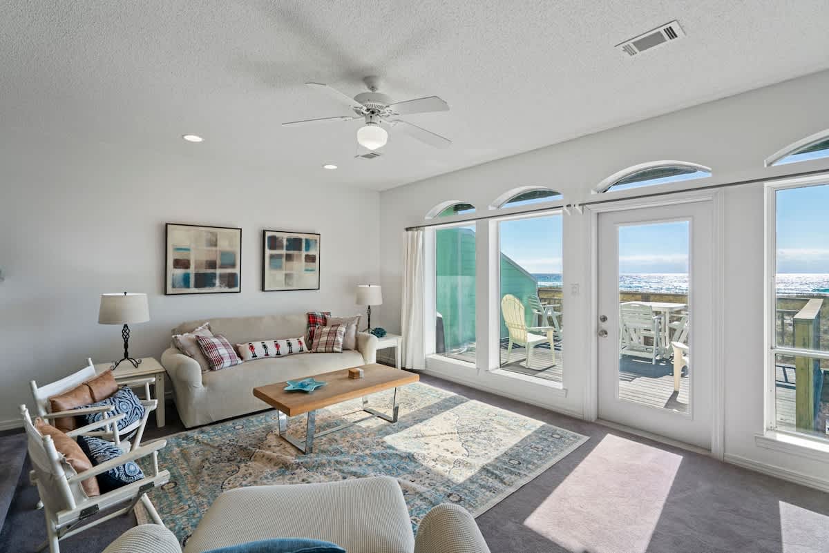 Bright living room with a light-colored sofa, two director-style chairs, and a wooden coffee table on a patterned rug, offering a clear view of the ocean through large windows and a glass door.