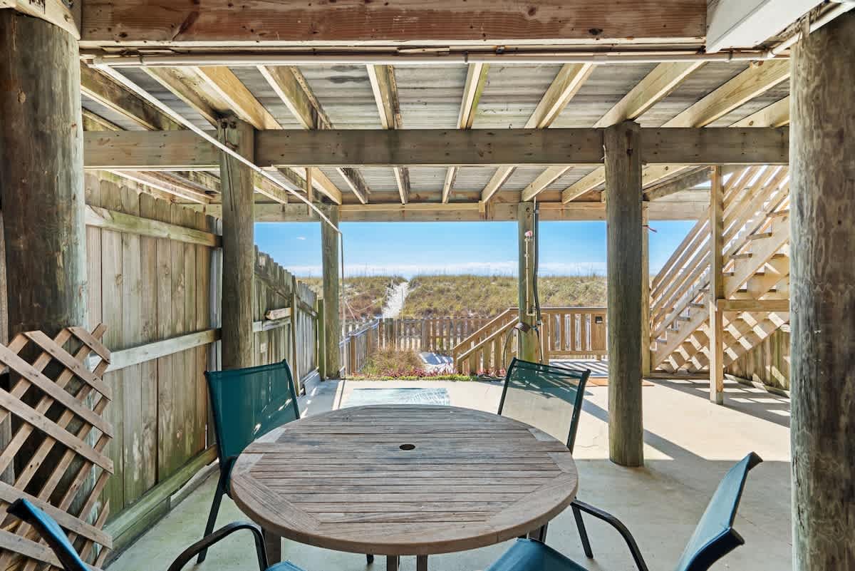 Covered outdoor patio area with a round wooden dining table and four chairs, offering a clear view of a sandy path leading to the beach and ocean. Wooden support beams are prominent overhead.