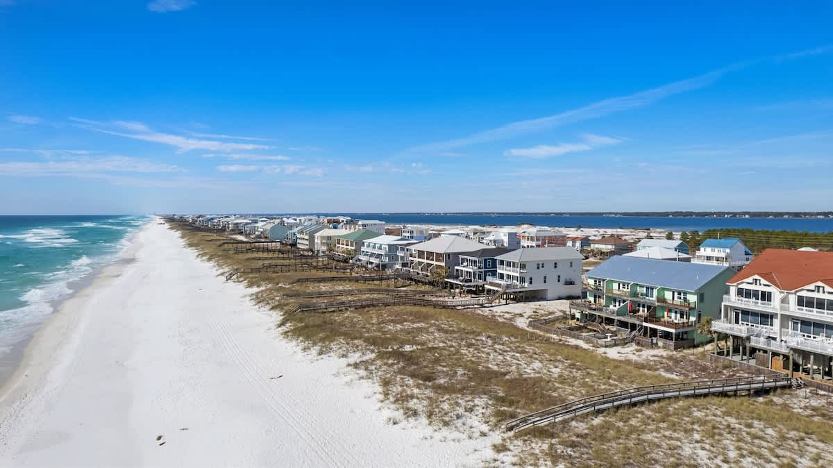 An expansive aerial view showcases a long stretch of pristine white sand beach bordering vibrant turquoise ocean waters. Numerous colorful beach houses with elevated foundations and wooden boardwalks are nestled among grassy dunes along the coastline.
