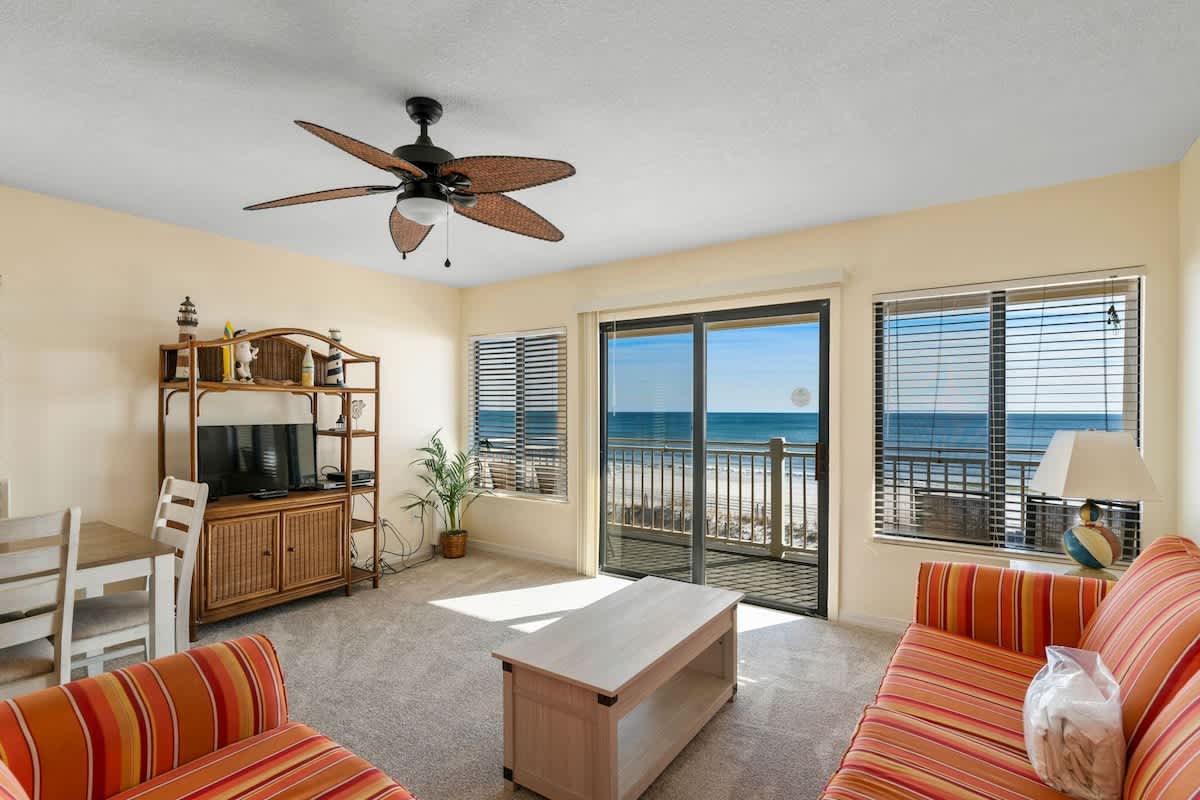 Bright living room with light yellow walls, carpeted floor, and an orange striped sofa and armchair, featuring a ceiling fan and a sliding glass door leading to a balcony with ocean views. A wooden media console with a TV and a small dining table are also visible.