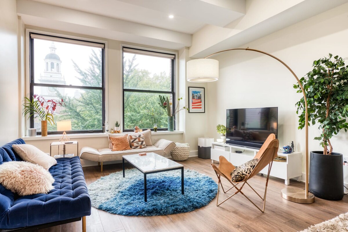 A bright living room featuring a blue tufted sofa, a beige chaise lounge, a marble coffee table, and a round blue rug, with large windows offering a view of a building and trees.