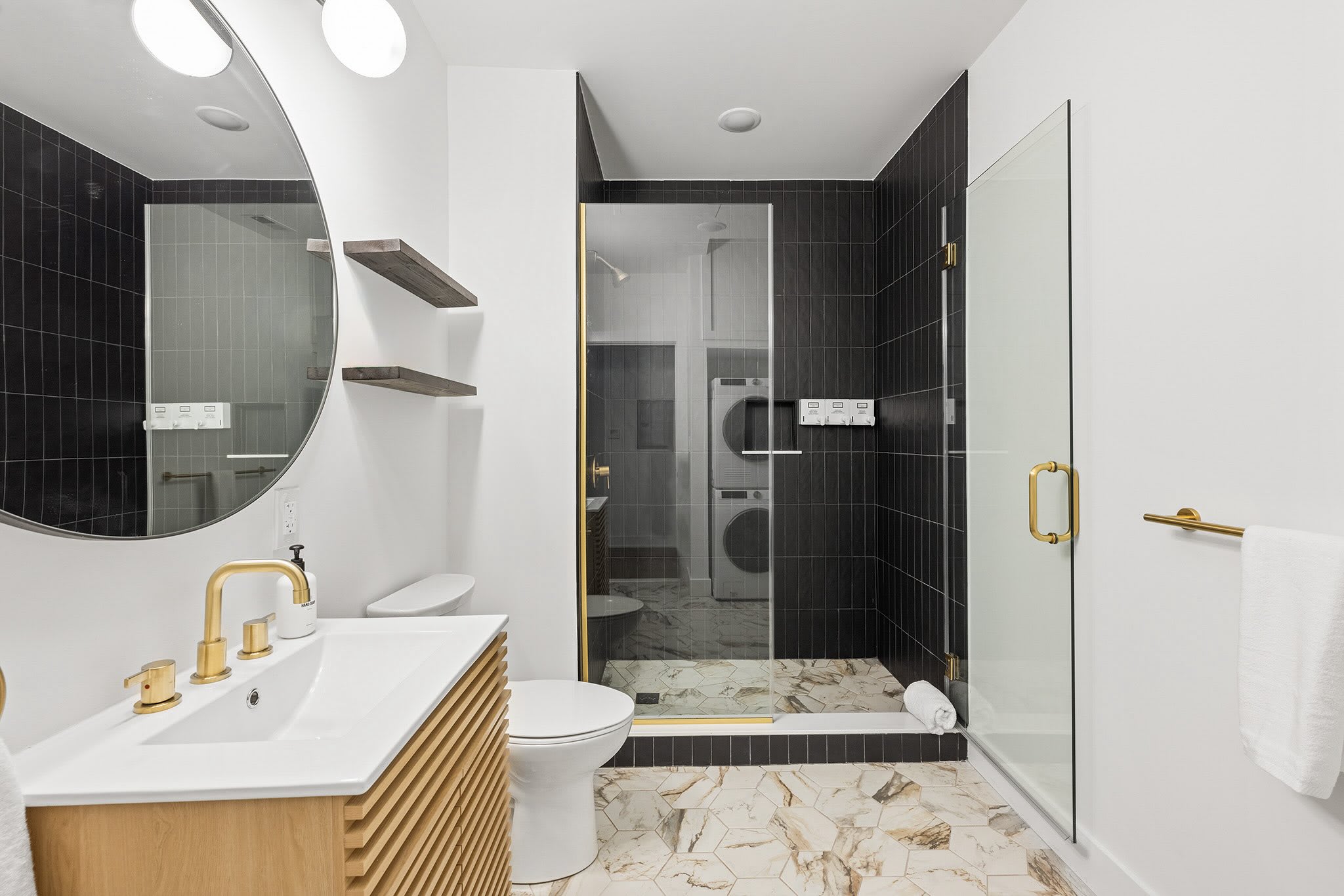 A stylish bathroom featuring a light wood vanity with a gold faucet, a large round mirror, and black subway tiles in the shower. The floor has geometric marble-patterned tiles, and a stacked washer/dryer is visible through the shower glass.