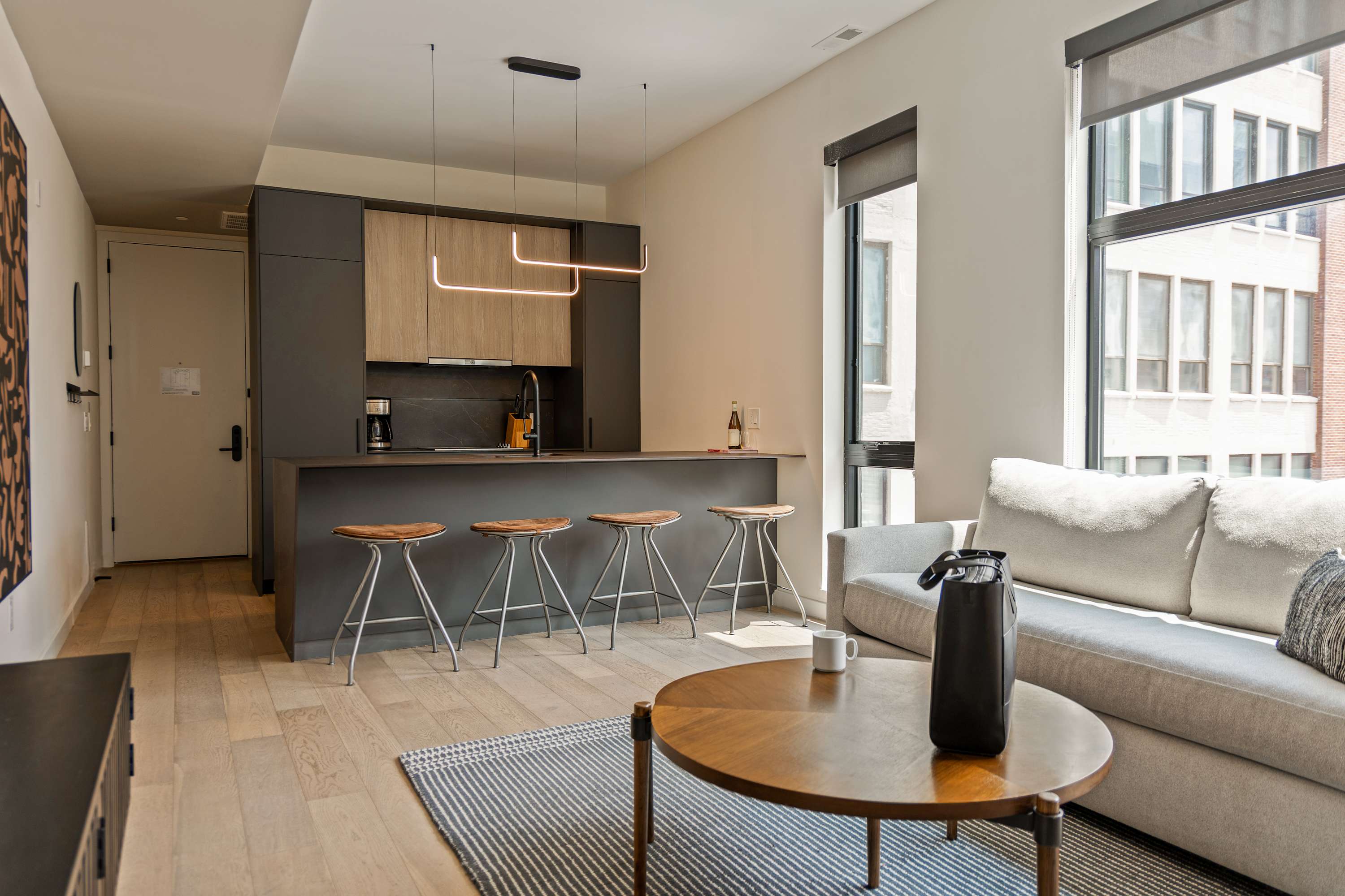 Open-concept living space featuring a modern kitchen with dark grey and light wood cabinetry, a breakfast bar with four wooden stools, and a light grey sofa in the living area. Large windows offer city views.