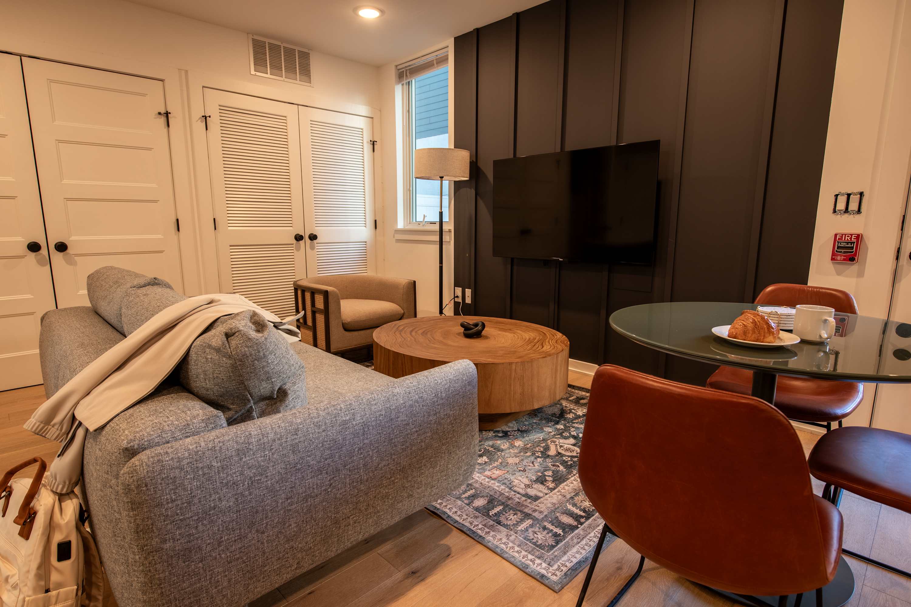A cozy living room with light wood floors and a patterned rug features a gray sofa with a throw blanket, a round wooden coffee table, and a dining area with a glass-top table and brown leather chairs.