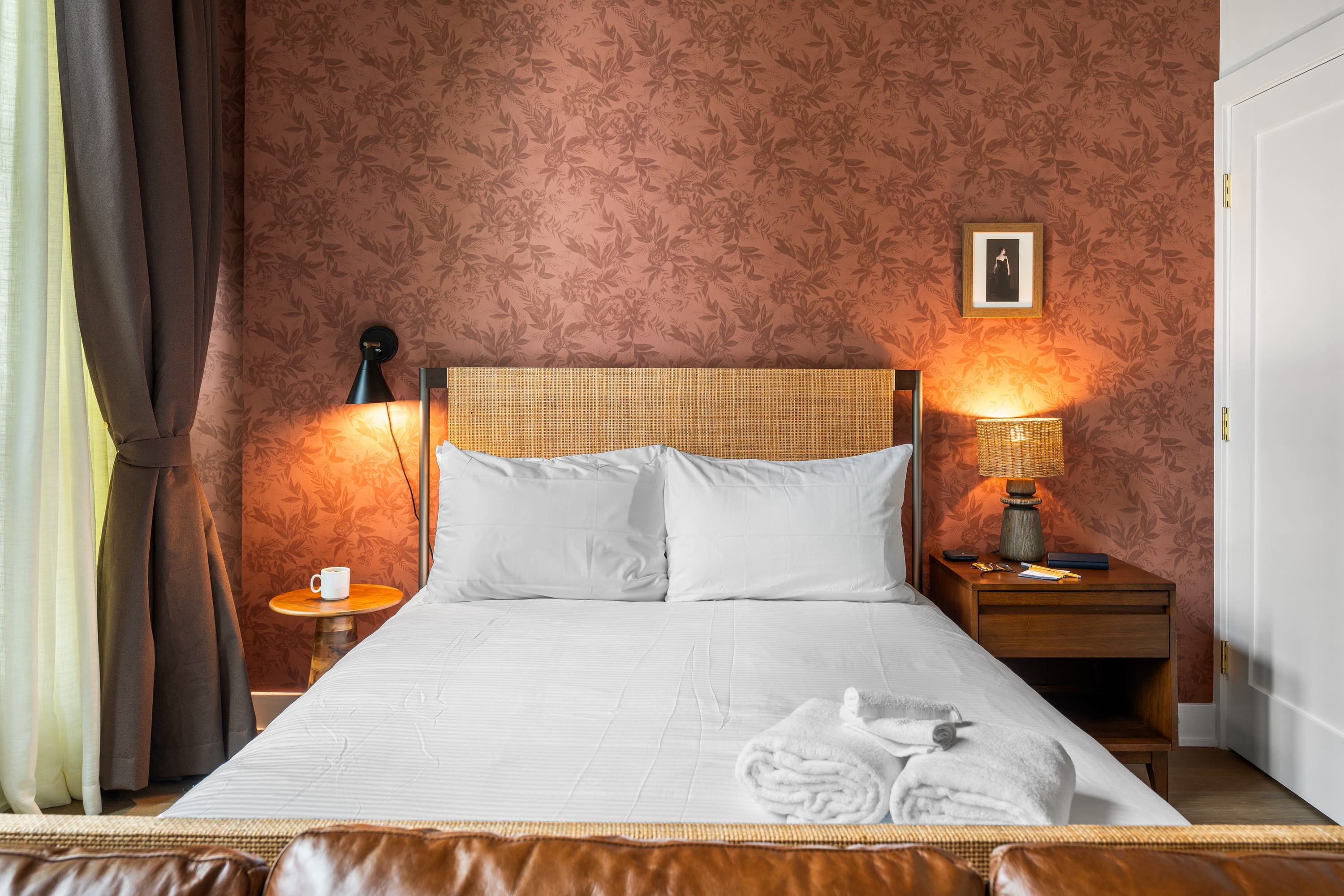 A cozy bedroom corner featuring a queen bed with a woven rattan headboard against a warm, floral-patterned accent wall. A wooden nightstand holds a wicker-shaded lamp, sunglasses, and a small framed picture.