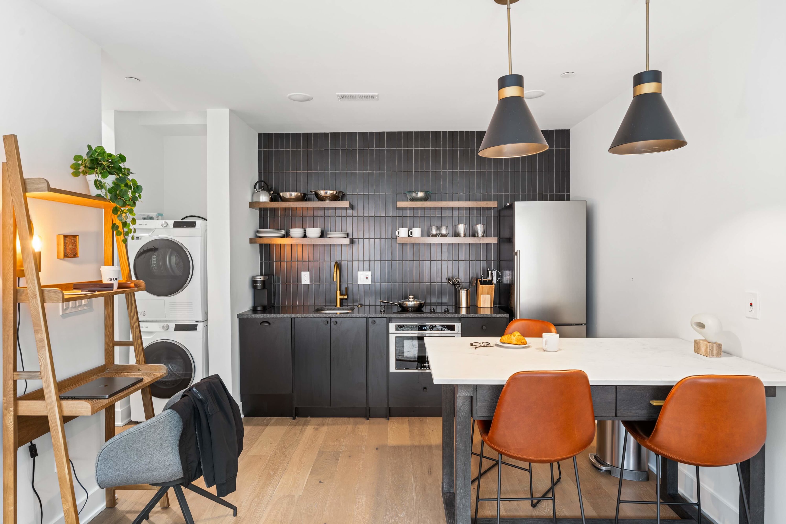 Modern kitchen with black flat-panel cabinetry, black subway tile backsplash, and stainless steel appliances including a stacked washer and dryer. A white quartz island with dark base and orange leather stools serves as a dining area.