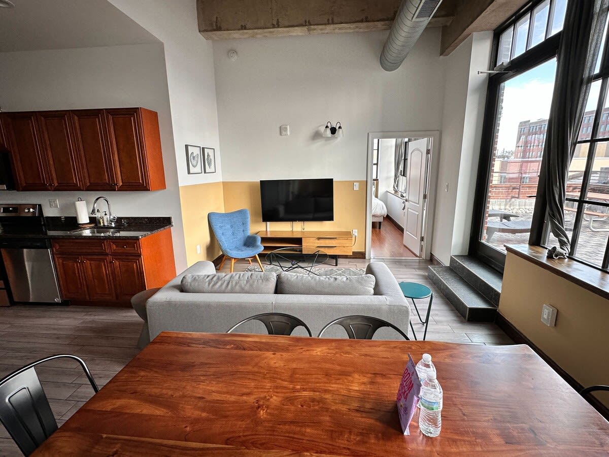 Open-concept living space with a light gray sofa, a wooden dining table with metal chairs, and a partial view of the kitchen, featuring a large window/door leading to an outdoor area.