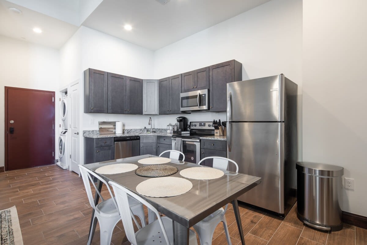 Well-equipped kitchen featuring dark gray cabinetry, granite countertops, and stainless steel appliances, with a metal dining table and white chairs in the foreground, and a stacked washer and dryer unit visible in a closet to the left.