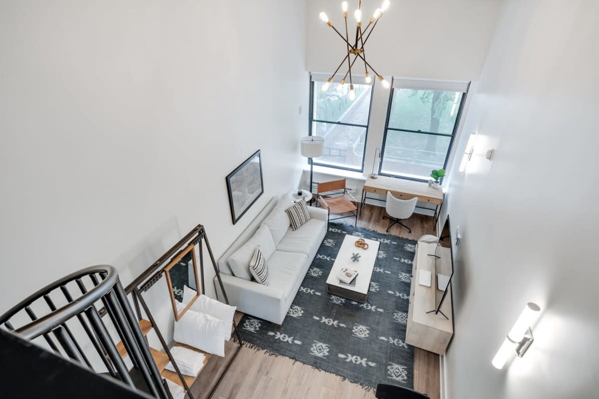 Overhead view of the living room from the loft, showcasing high ceilings, large windows, a light-colored sofa, and a desk area with a chair. A patterned rug covers the light wood floor, and a modern chandelier hangs from above.
