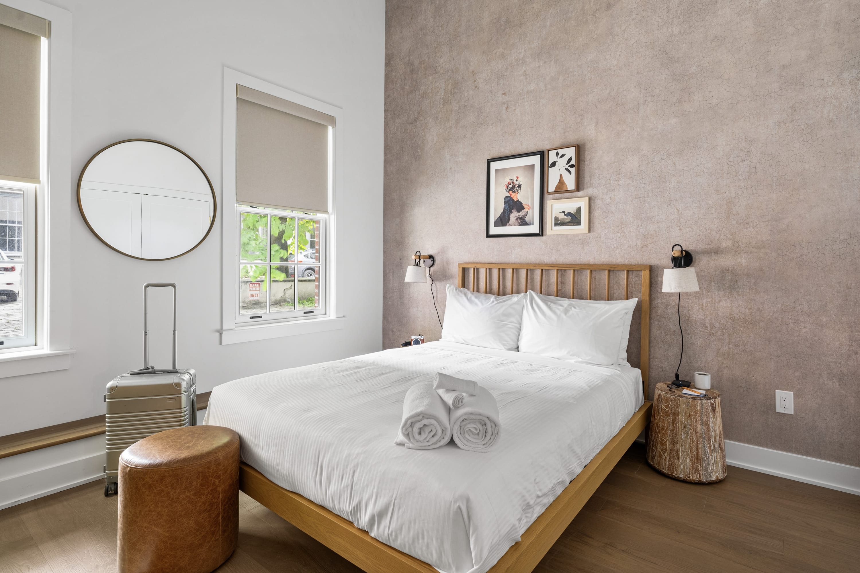 Bright bedroom with a light wood bed frame, crisp white bedding, a round wall mirror, and a textured beige accent wall, illuminated by natural light from two windows.
