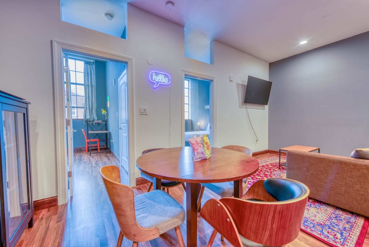 A vibrant living and dining area featuring a round wooden dining table with four mid-century modern chairs, a "hello" neon sign, and a wall-mounted TV above a brown sofa on a red patterned rug.