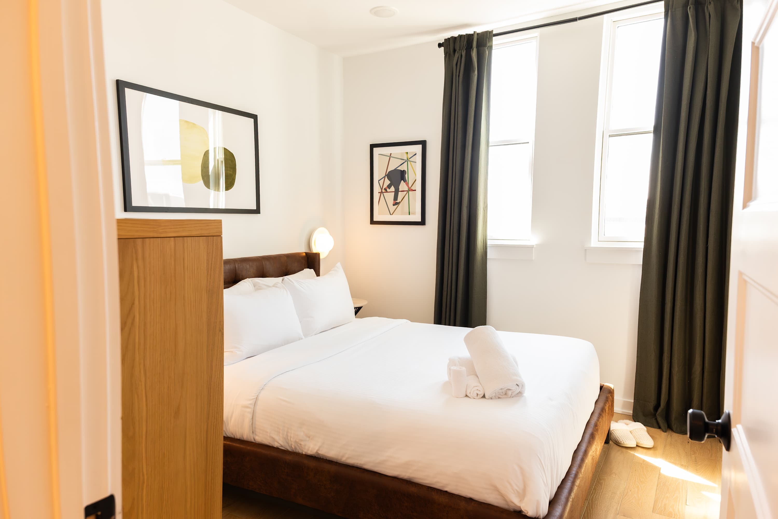 A bright bedroom featuring a brown upholstered bed with white linens and two windows draped with dark green curtains. Abstract artwork adorns the white walls, and a wooden cabinet is partially visible on the left.