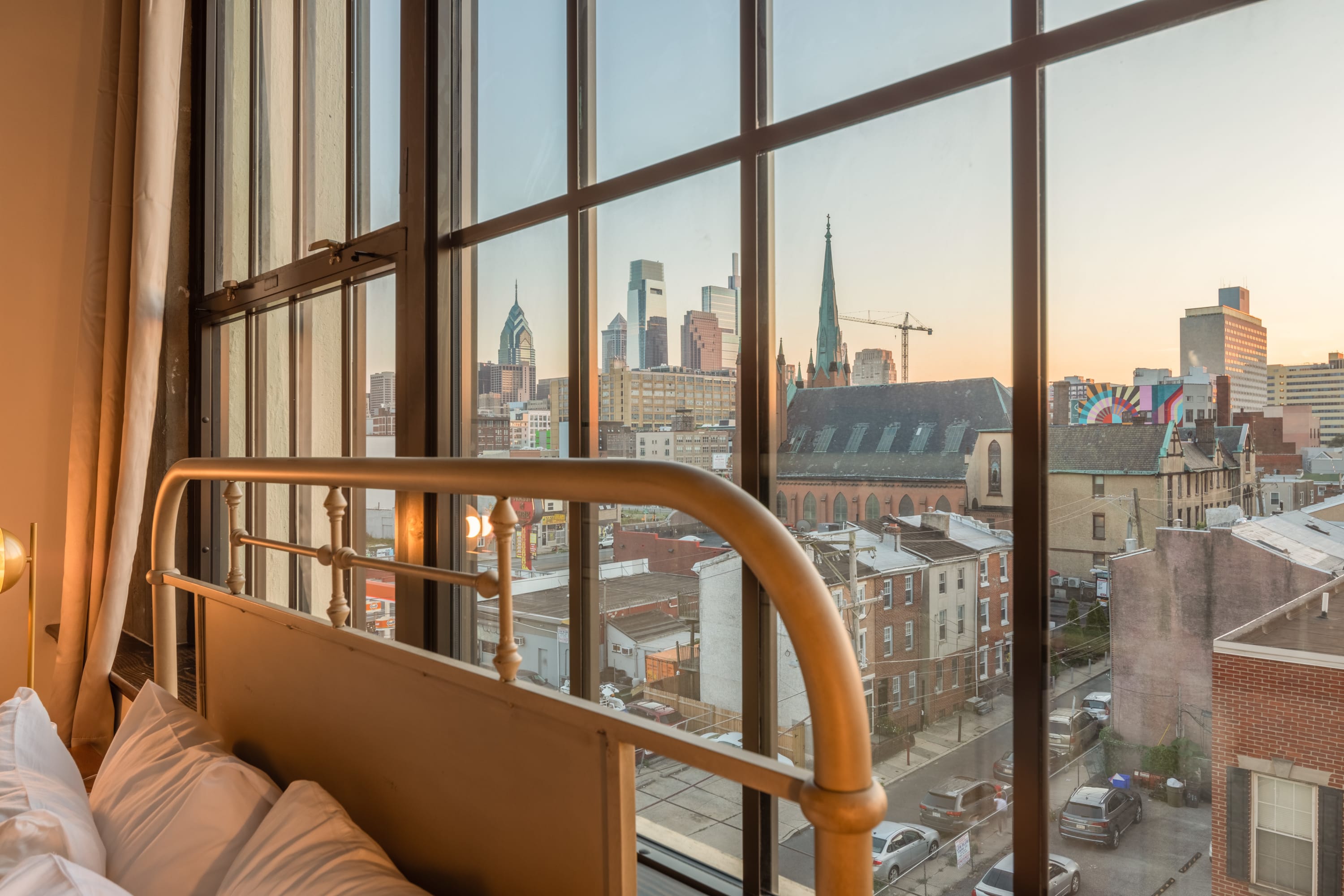 A close-up view from a large industrial-style window showcasing a city skyline with various buildings, including a prominent church steeple and modern skyscrapers, under a soft evening sky. A metal bed frame is visible in the foreground.