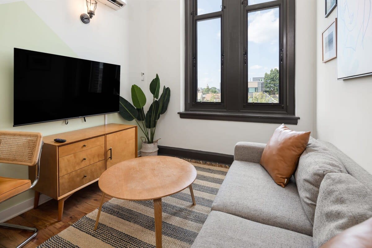 A modern living space with a light gray sofa, a round wooden coffee table, and a striped area rug. A large flat-screen TV is mounted above a wooden console, next to a tall green plant, with a tall window providing natural light.