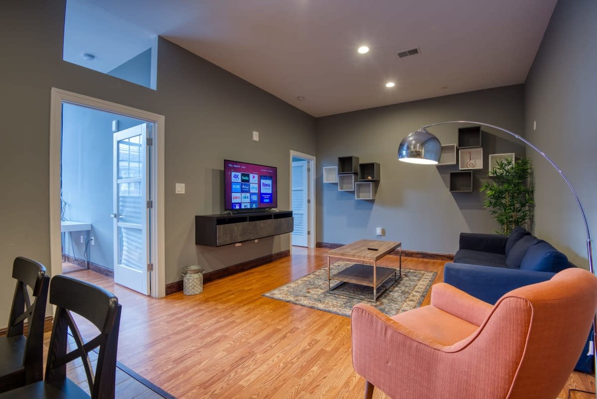 A spacious living room with light hardwood floors and dark gray walls, featuring a large wall-mounted TV above a floating media console. A dark blue sofa and an orange armchair are arranged around a wooden coffee table on a patterned rug, with decorative cube shelves adorning the wall.