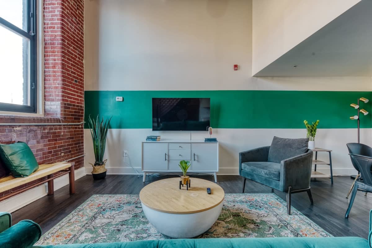 A bright living room featuring a teal velvet sofa, a white media console with a TV, and a gray armchair, all arranged on a patterned area rug. A bold green stripe painted across the white wall adds a modern accent, complementing the exposed brick wall and large windows.