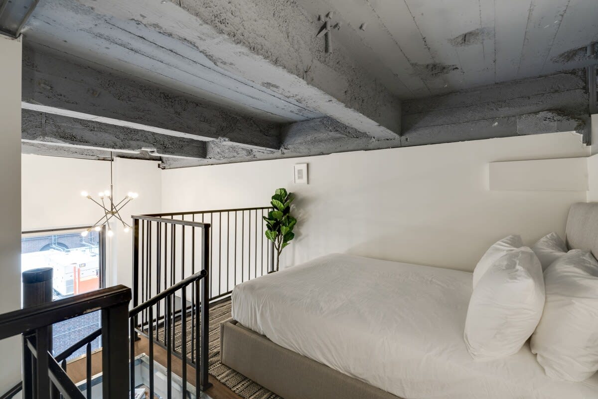 A cozy loft bedroom showcases a bed with white bedding, a black metal railing overlooking the lower level, and an industrial-style exposed concrete ceiling with dark beams. A modern chandelier and a small plant add decorative touches.