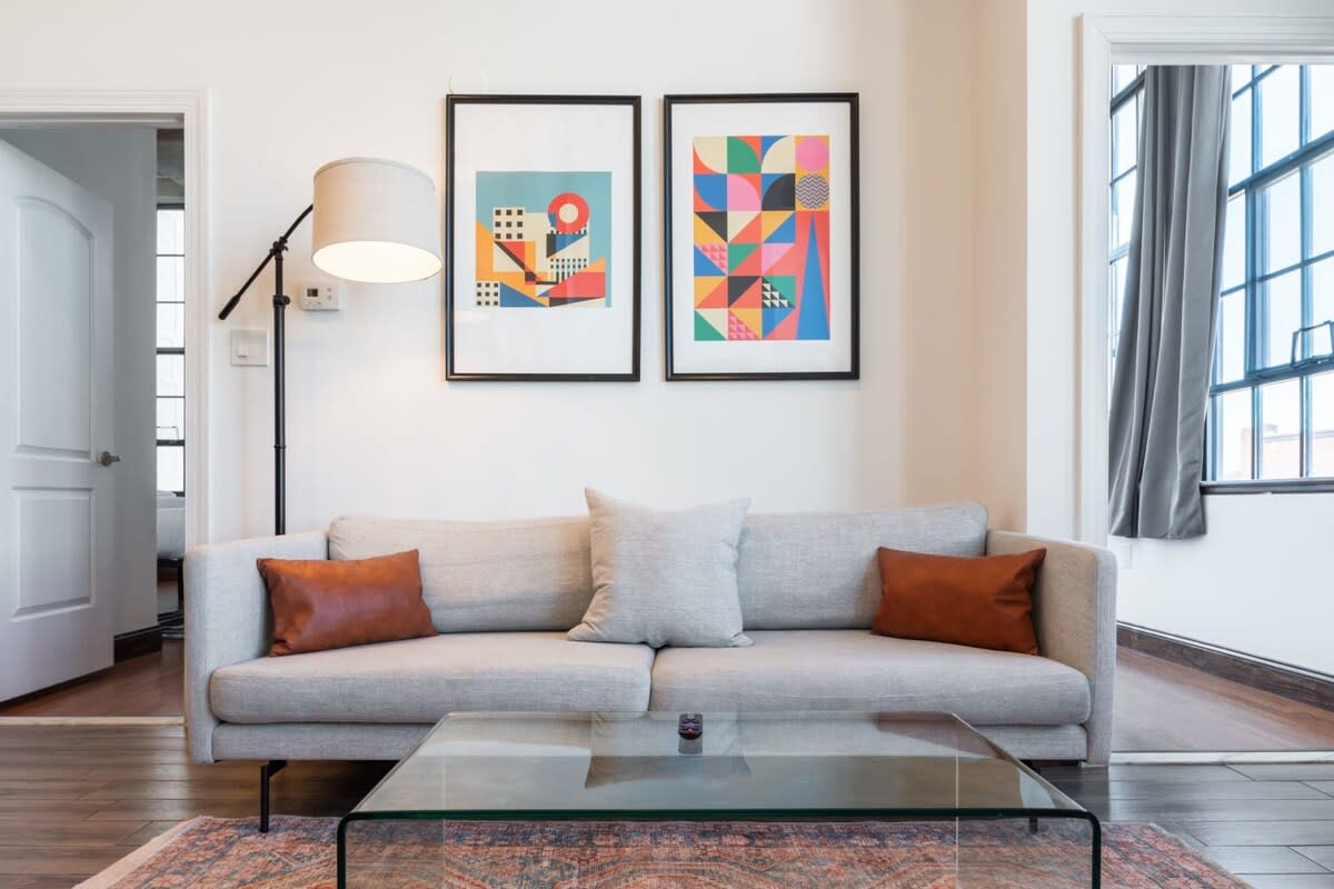 Cozy living room featuring a light gray sofa with brown accent pillows, a glass coffee table, and a colorful patterned rug, flanked by two abstract art prints on the wall. A floor lamp provides ambient lighting.