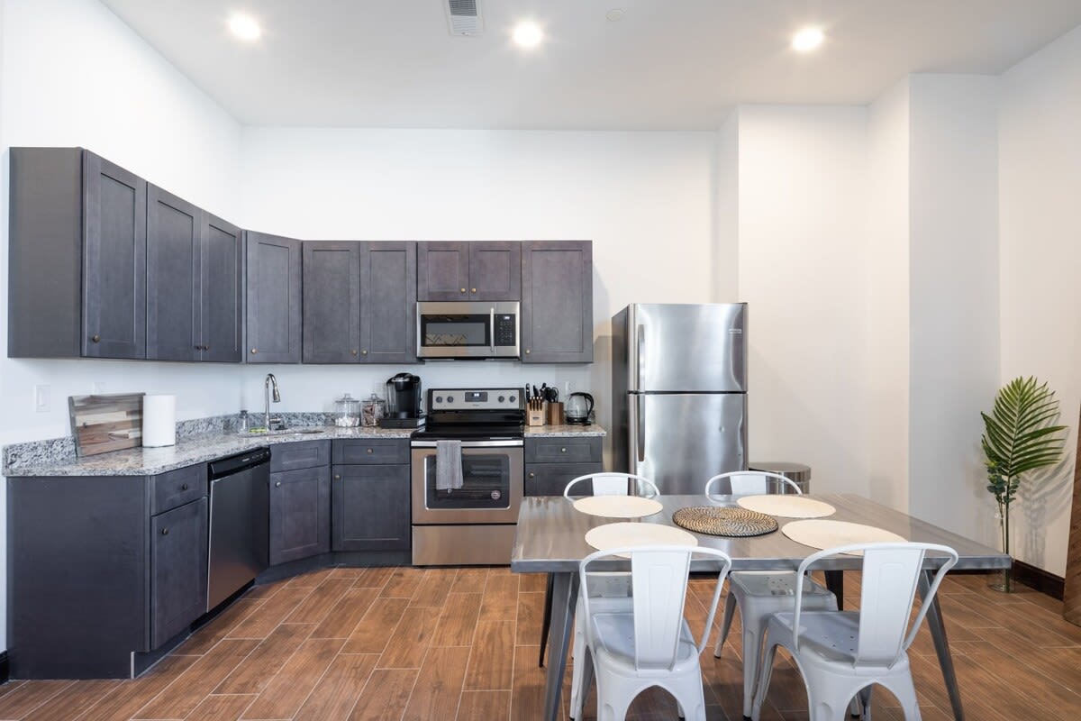 Modern kitchen with dark gray shaker-style cabinets, speckled granite countertops, and stainless steel appliances including a refrigerator, microwave, dishwasher, and range, adjacent to a metal dining table with white chairs.