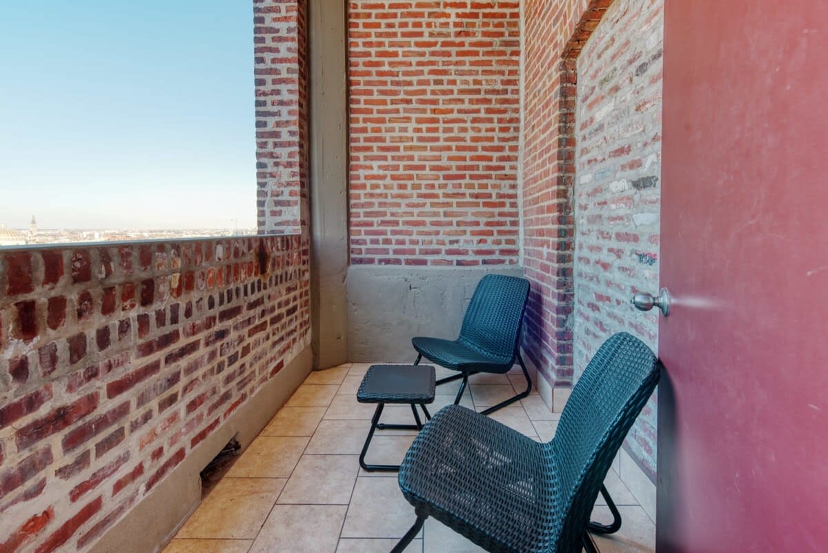 Small outdoor balcony with red brick walls and light-colored tiled flooring, furnished with two black chairs and a small black table, offering a glimpse of the city skyline.