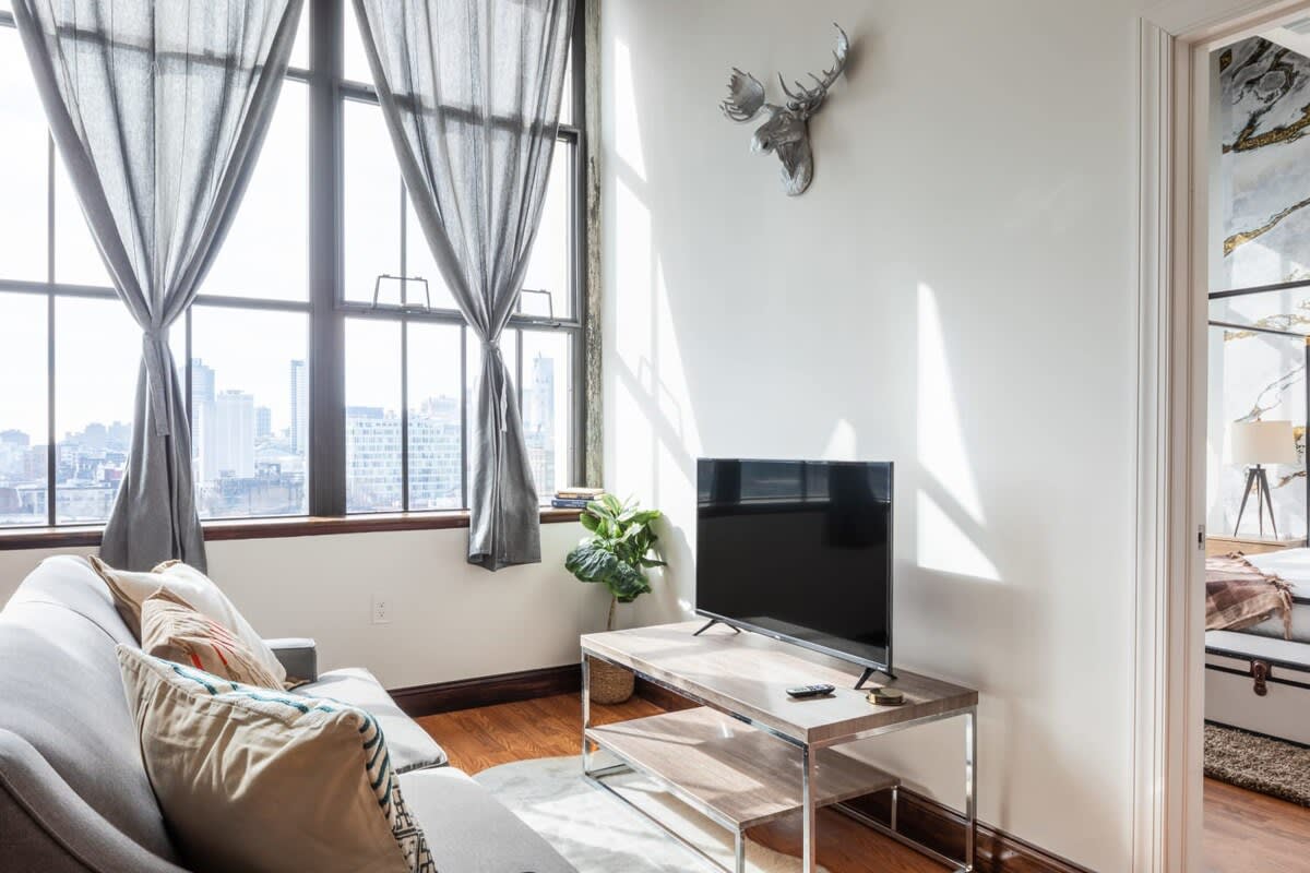 Cozy living room with a light grey sofa adorned with decorative pillows, facing a flat-screen TV on a minimalist wooden stand. Large windows provide a bright atmosphere and city views, complemented by a decorative deer head on the wall.