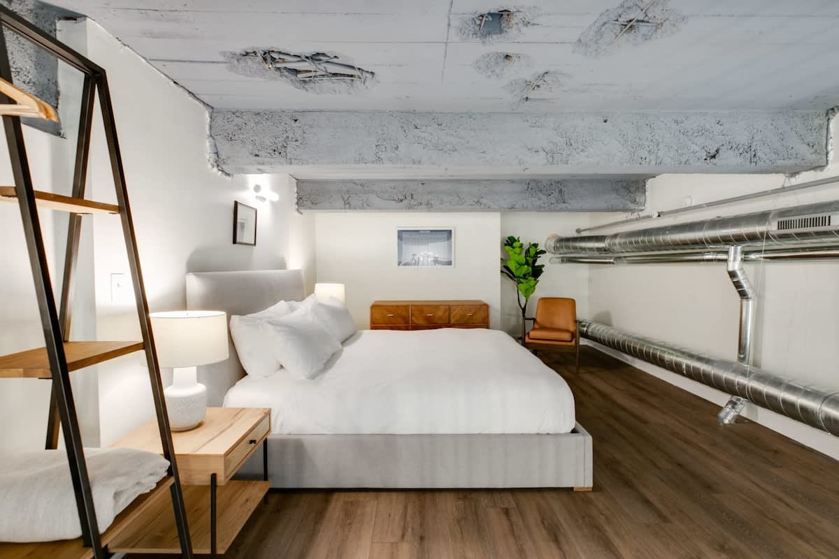 A minimalist bedroom located in a loft space, featuring a queen-sized bed with white bedding, a wooden nightstand, and an industrial-style ceiling with exposed concrete and ductwork. A brown leather chair and a plant add warmth.