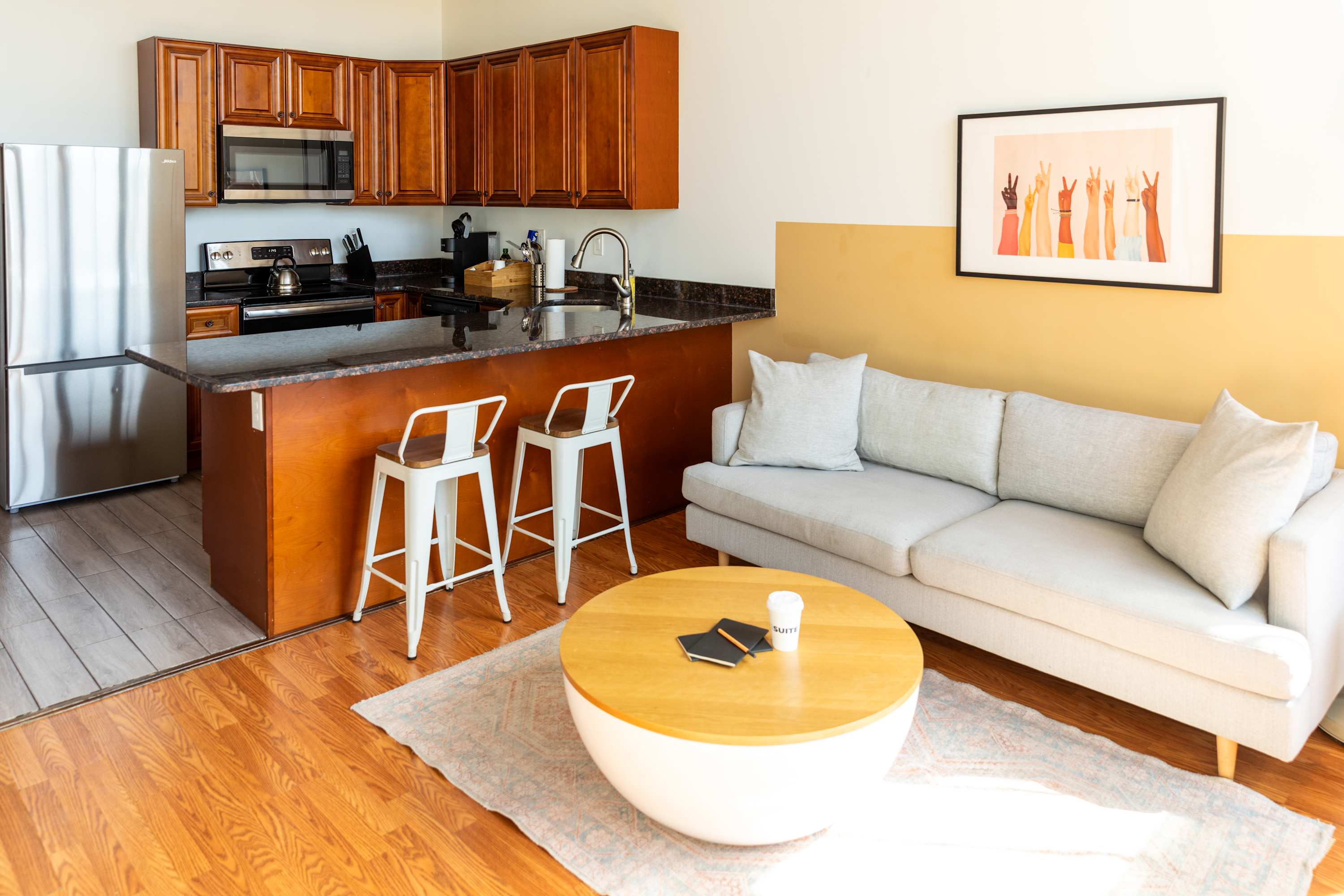 Open-concept kitchen with warm wood cabinetry, dark granite countertops, and stainless steel appliances, including a refrigerator and microwave. Adjacent living area features a light-colored sofa and a round coffee table.