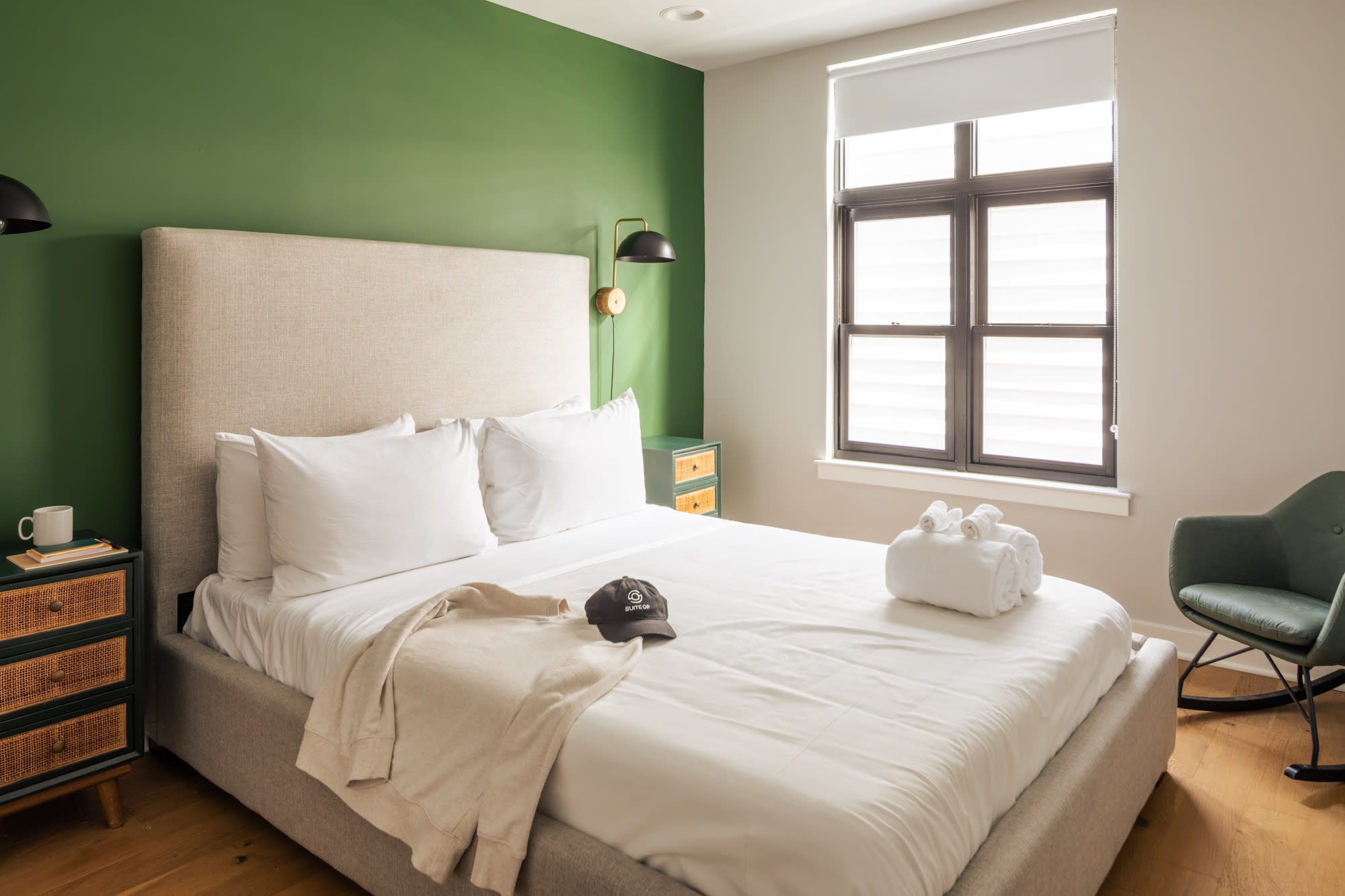 A cozy bedroom with a prominent green accent wall behind a large bed with a light beige upholstered headboard, white bedding, and two dark green nightstands with woven drawers. Two black and gold wall sconces flank the bed.