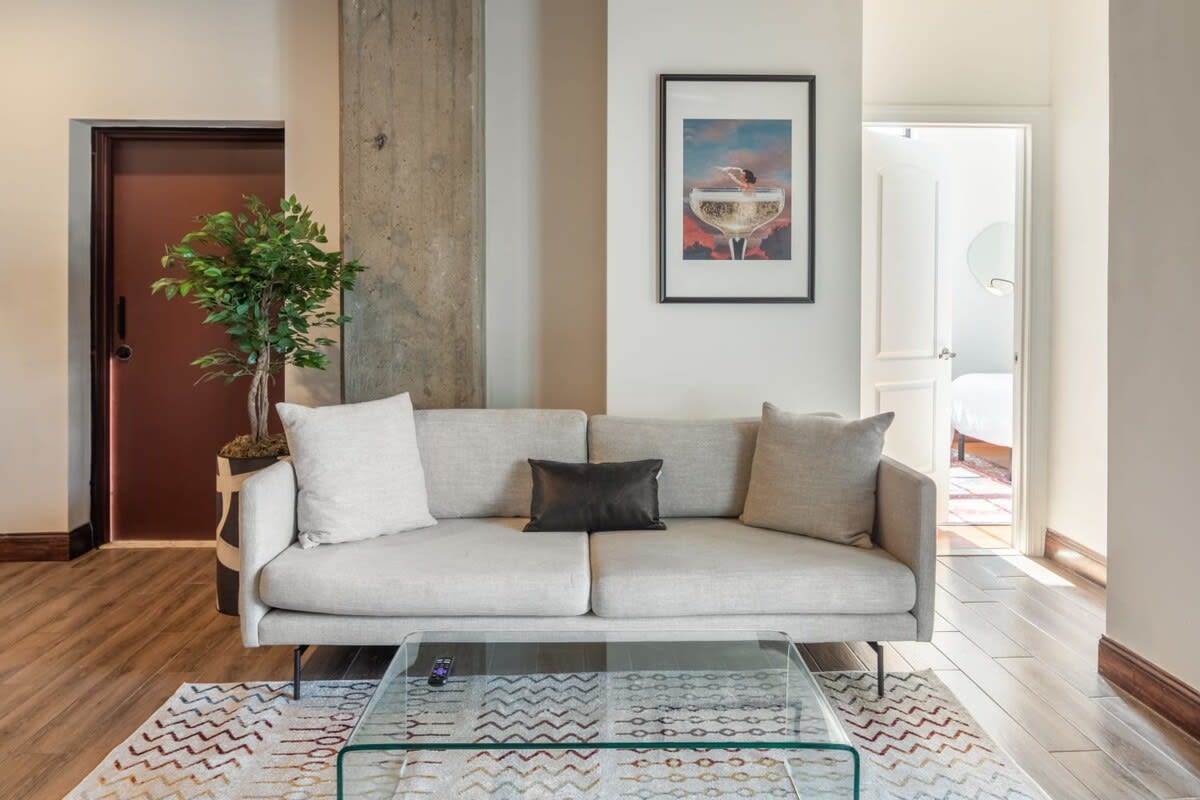 A comfortable living room area featuring a light grey sofa with throw pillows, a clear glass coffee table, and a geometric patterned area rug on wood-look flooring. A tall potted plant stands beside a concrete pillar, and a framed art piece hangs on the wall, adding a touch of decor.