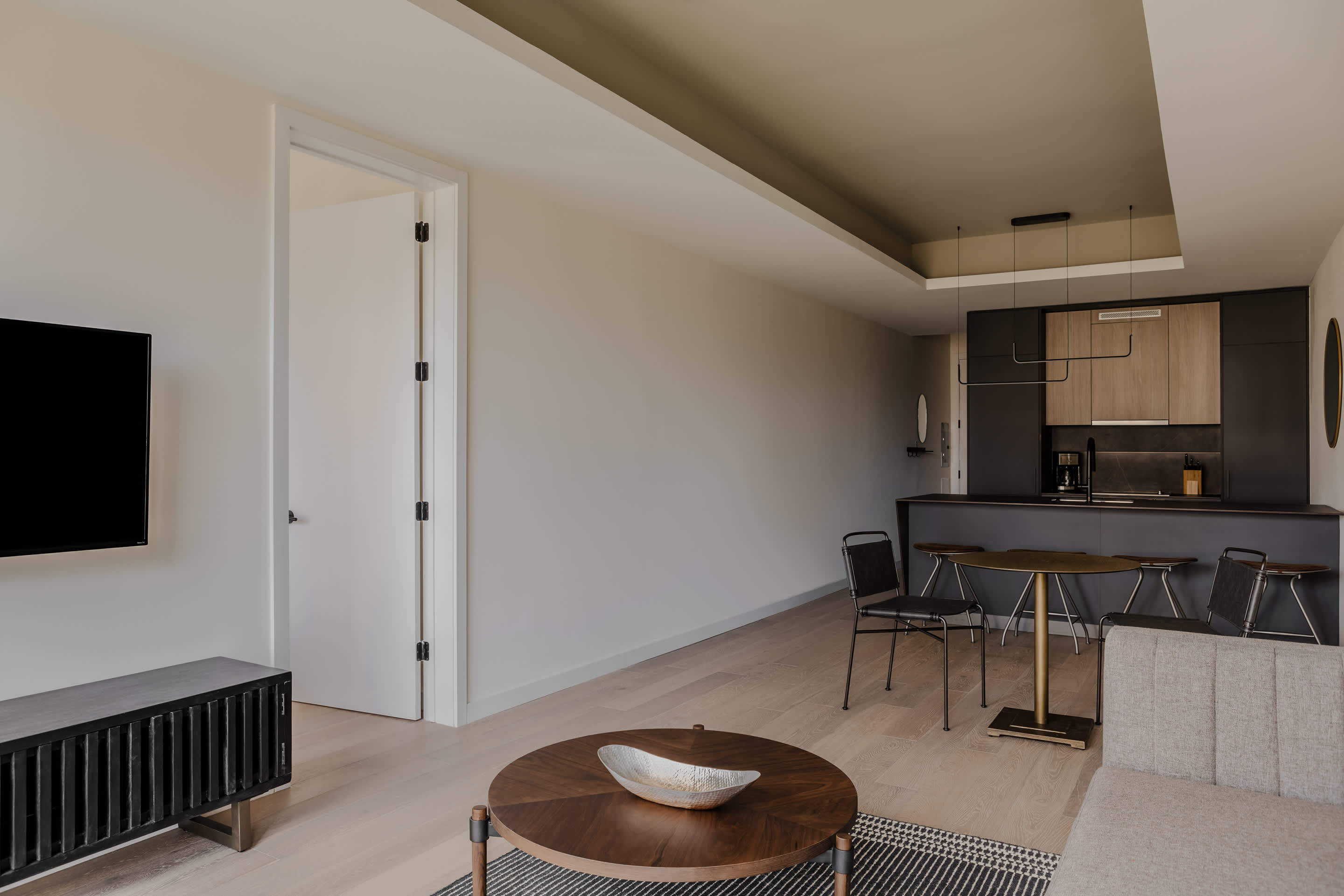 Open-concept living area with a black TV console and a round dark wood coffee table, leading into a modern kitchen and dining space with light wood flooring.