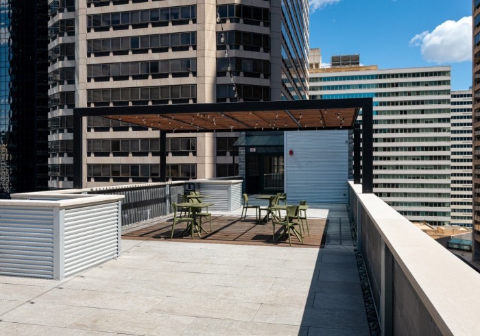 An expansive rooftop deck offering city views, featuring a shaded pergola with string lights over a seating area with green metal chairs and tables. The deck has light-colored paving stones and planters.
