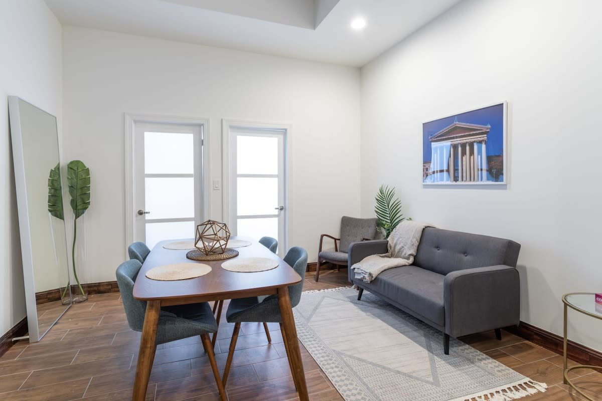A bright living and dining area with white walls and wood-look tile flooring, featuring a wooden dining table with blue chairs and a gray sofa with an accent chair. A large mirror and plant add decorative touches.