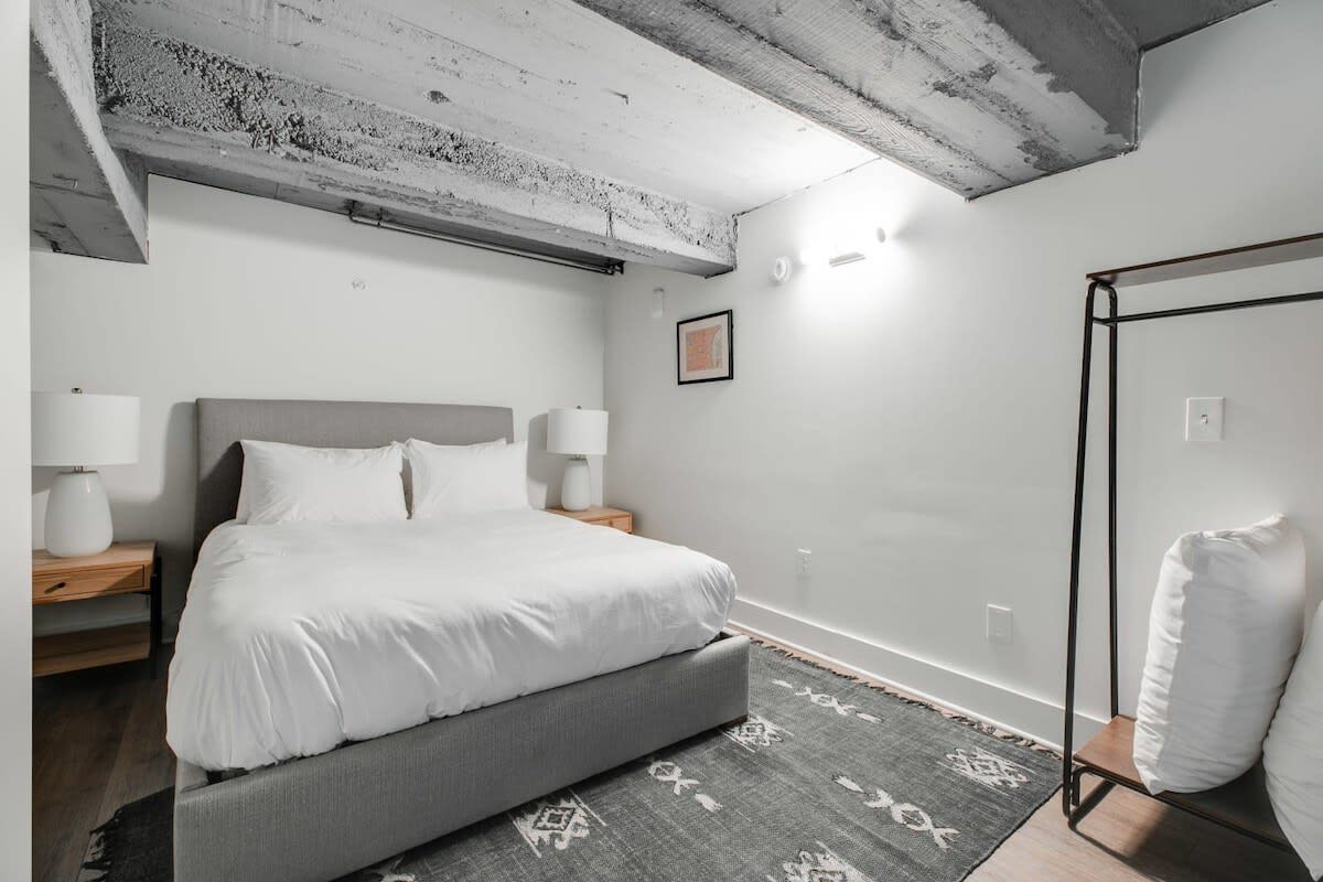 A cozy bedroom with a queen-sized bed featuring a grey upholstered headboard and crisp white bedding. Two wooden nightstands with lamps flank the bed, and a patterned grey rug covers the hardwood floor. Exposed concrete beams are visible on the ceiling.