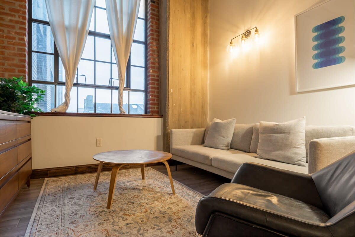 Bright living area with a light gray sofa, a black leather lounge chair, and a round wooden coffee table, illuminated by large industrial-style windows with white sheer curtains and a brick wall accent.