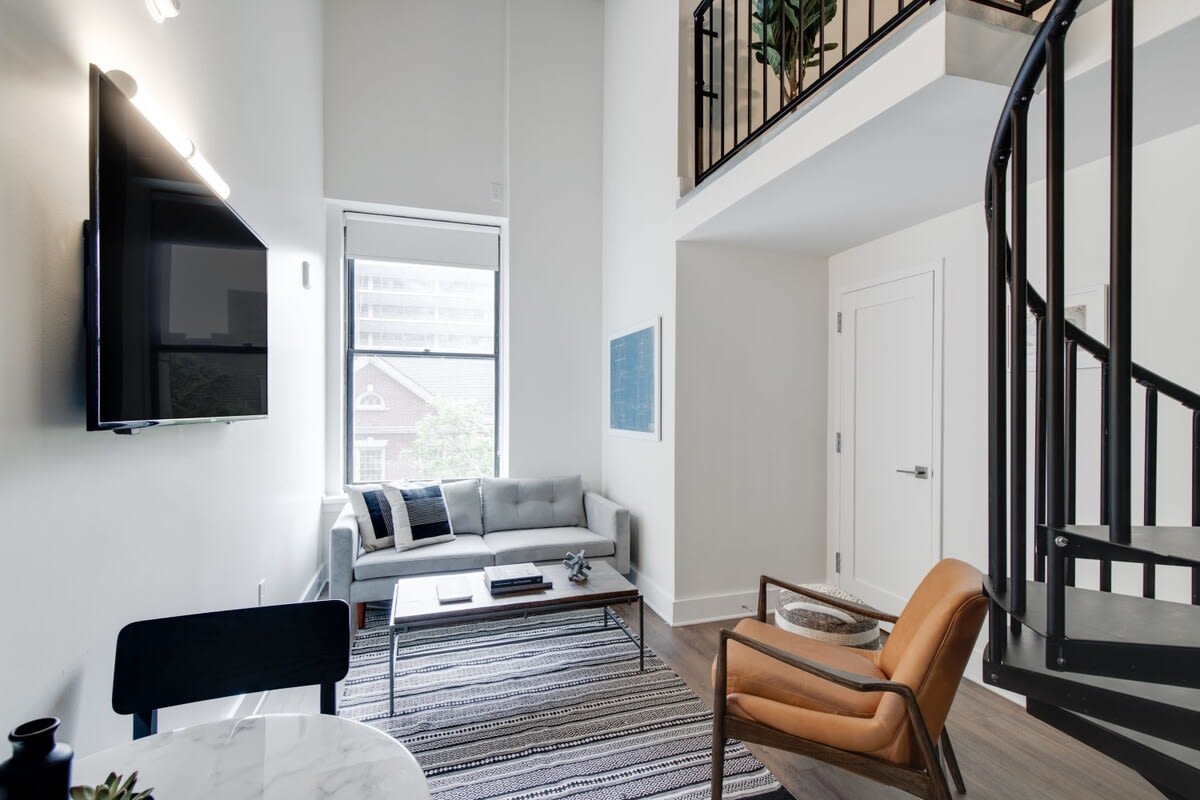 Modern living room with a light grey sofa, a dark wood coffee table on a striped rug, a wall-mounted TV, and a large window providing natural light, adjacent to a black spiral staircase.