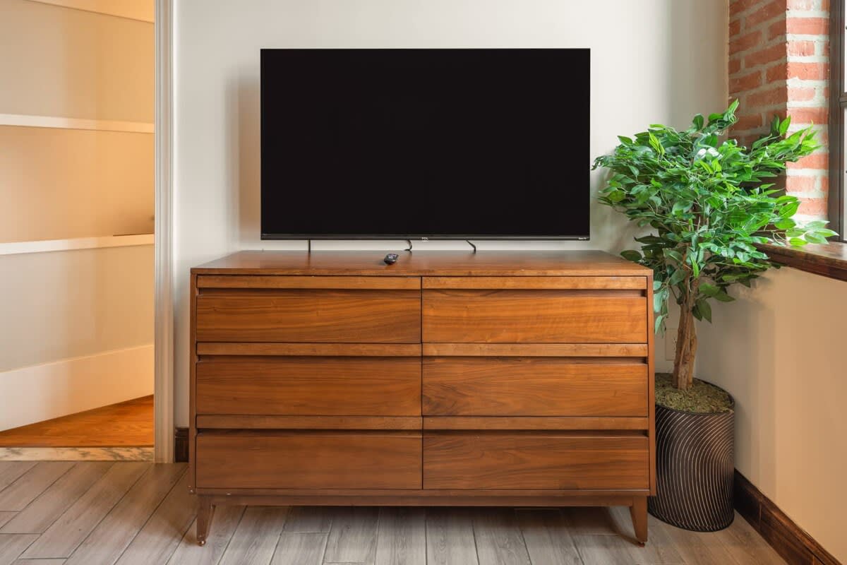 Detail shot of the living room featuring a large flat-screen TV resting on a mid-century modern wooden dresser with six drawers, accompanied by a tall potted artificial plant.