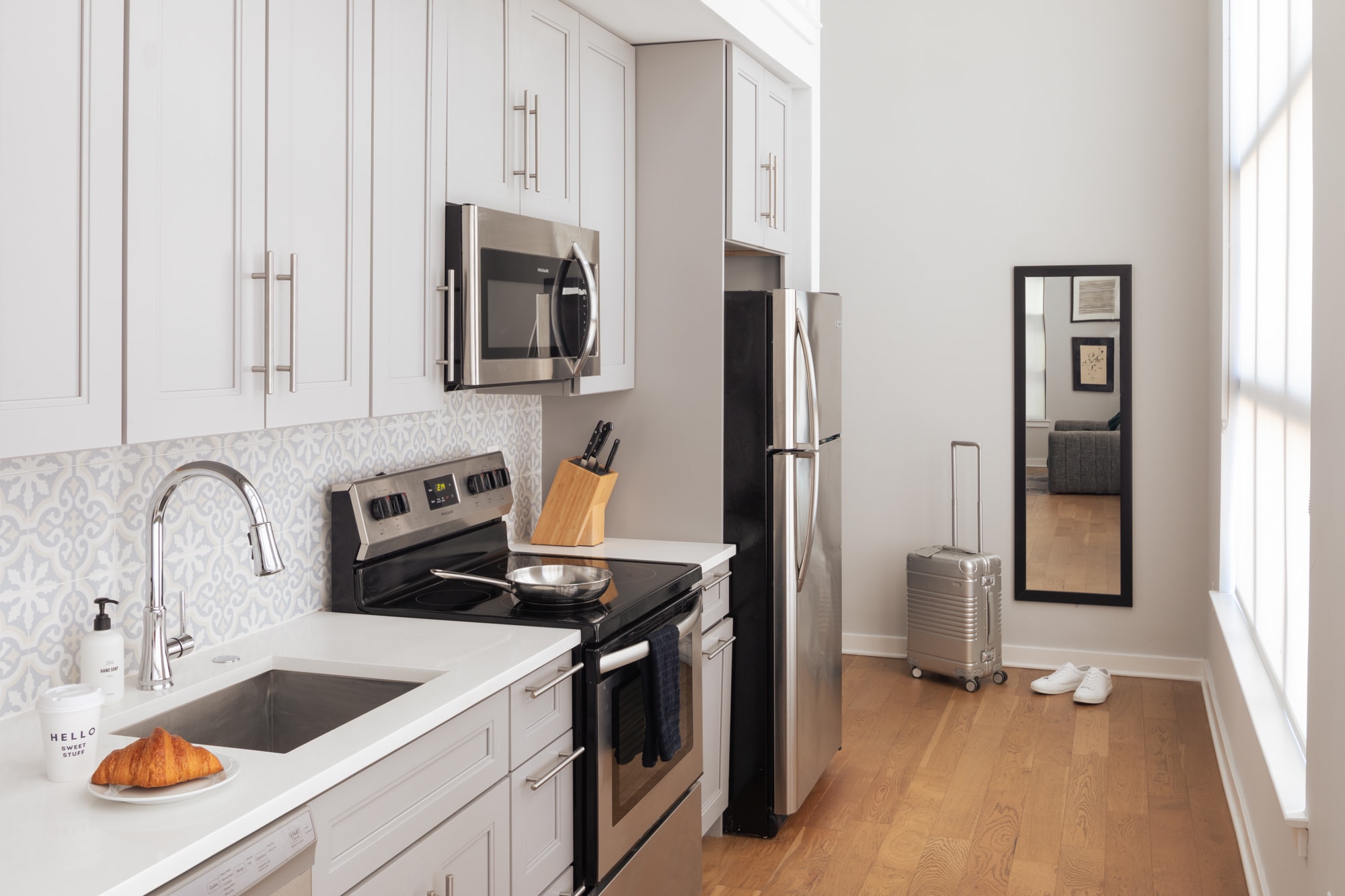 A modern kitchen with light gray shaker cabinets, stainless steel appliances including a refrigerator, microwave, and range, white countertops, and a light blue patterned backsplash. A silver suitcase and white shoes are visible near a full-length mirror.