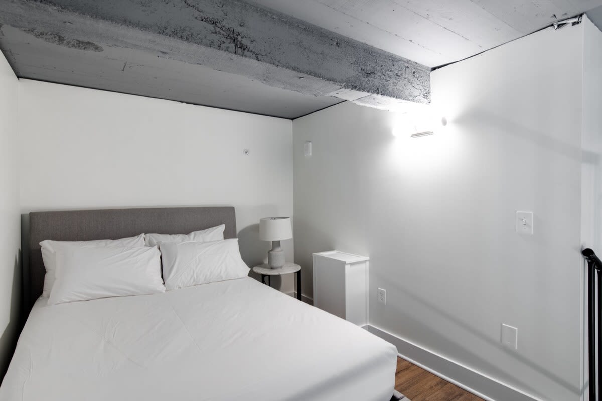 Loft bedroom featuring a queen-sized bed with a grey upholstered headboard, white bedding, and exposed grey concrete ceiling beams, with a small nightstand and lamp.