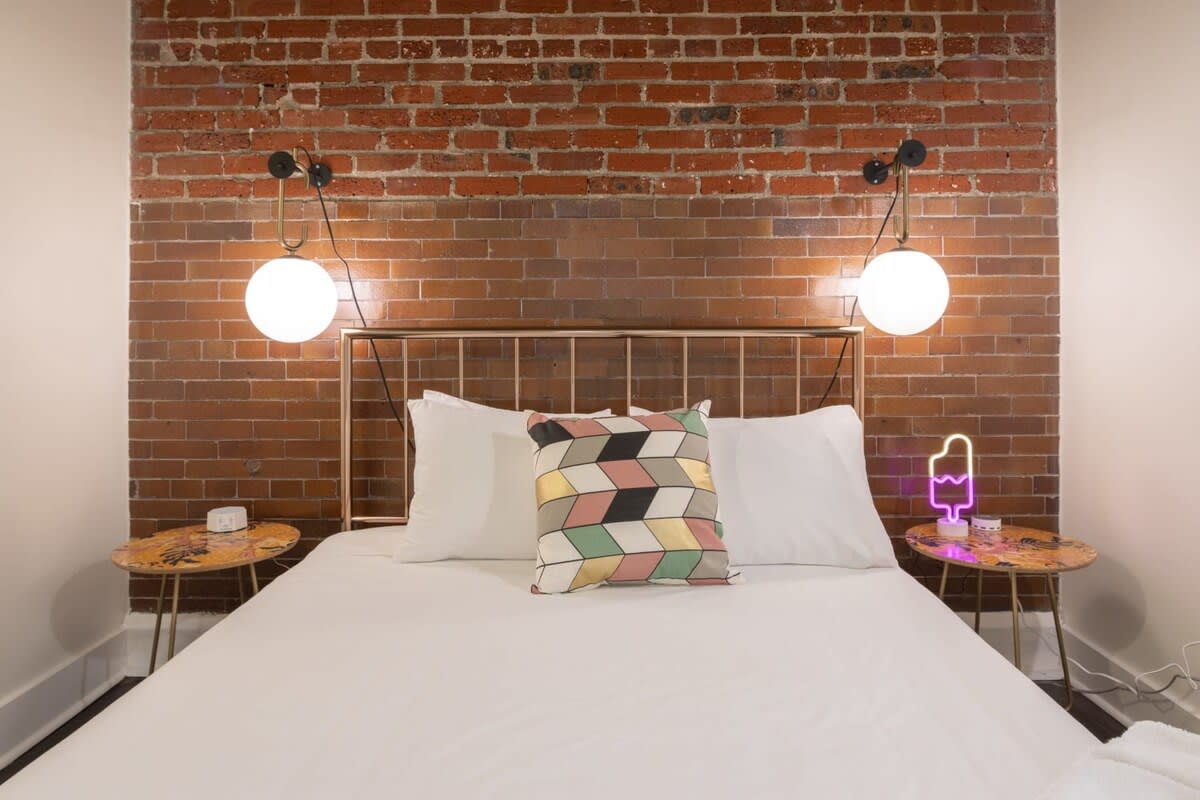 Stylish bedroom with a white bed, two unique round side tables, and modern wall-mounted globe lights against an exposed brick accent wall. A colorful geometric pillow adds a pop of color.